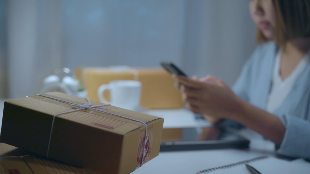 Smart Asian young entrepreneur business woman owner of SME online checking product on stock and save to mobile phone working late in night at home.